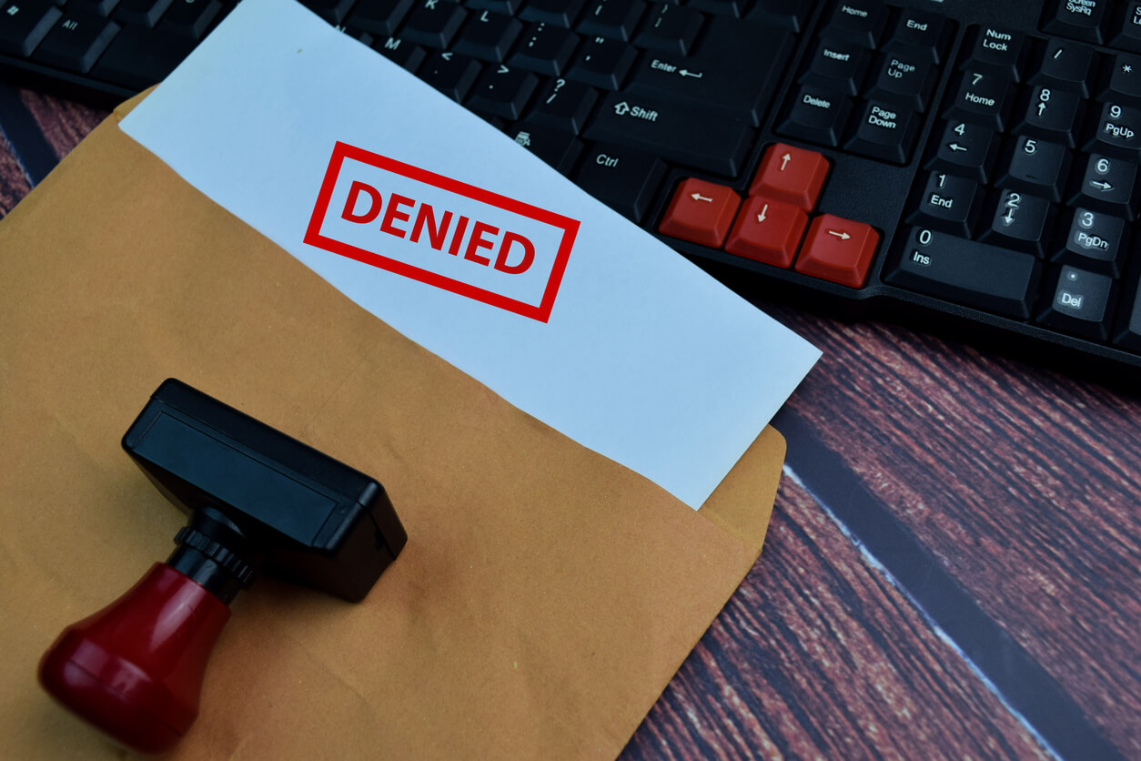 Document stamped 'DENIED' on a white paper partially inside a brown envelope, with a rubber stamp and a computer keyboard in the background.
